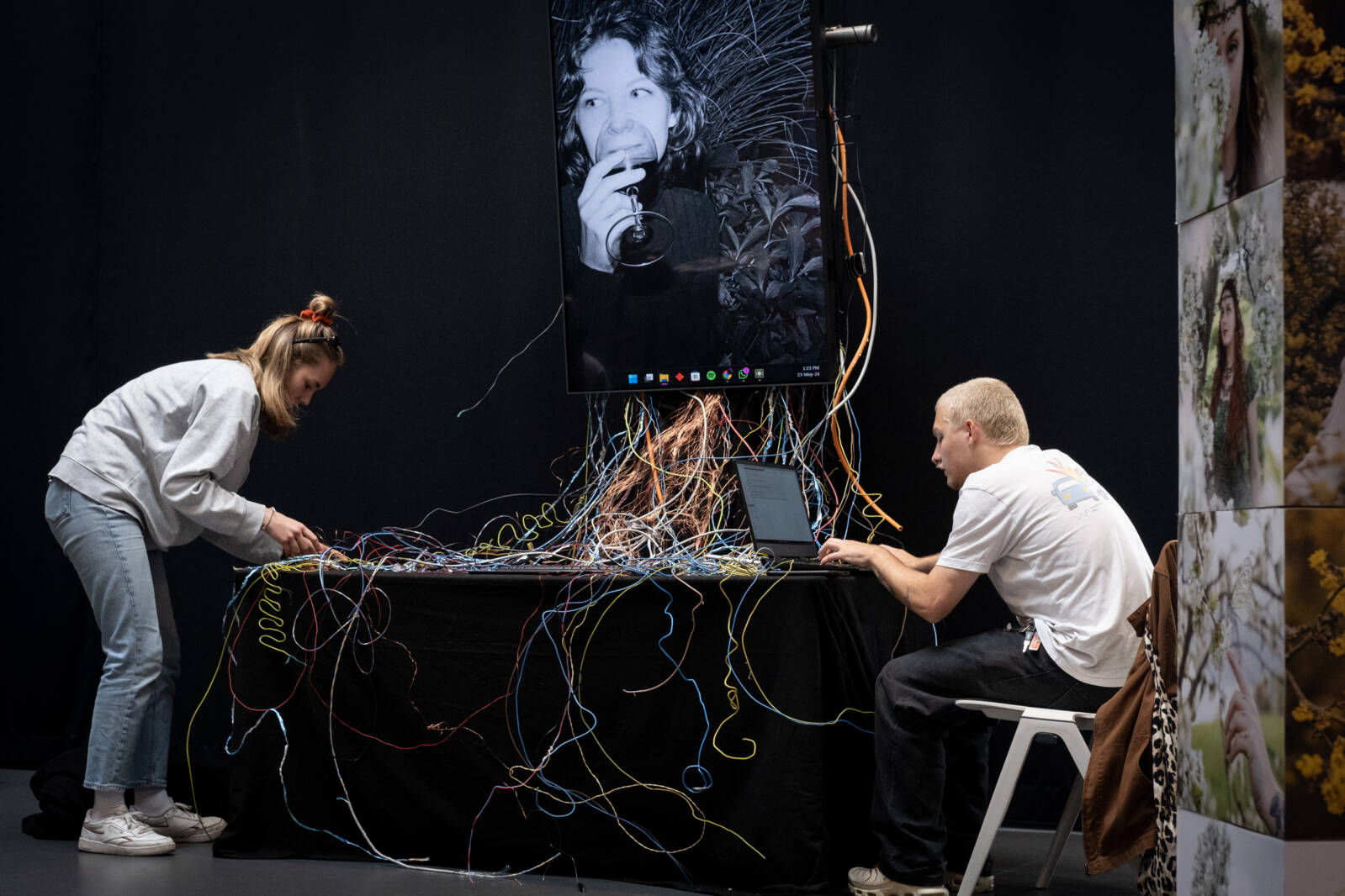 Zwei Personen arbeiten an einer Kunst- und Technologieinstallation mit bunten Kabeln, die zu einem großen Bildschirm mit einem Schwarz-Weiß-Porträt führen. Eine Person richtet die Kabel, während die andere am Laptop arbeitet.